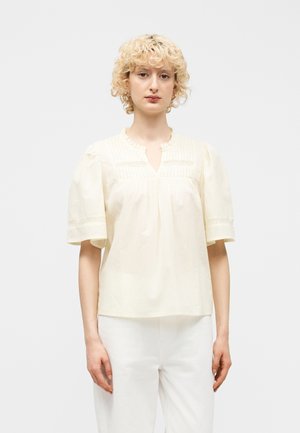 Kobieta o krótkich kręconych blond włosach, ubrana w kremową bluzkę z krótkim rękawem z koronkowymi detalami i białe spodnie, stojąca na jednolitym tle.