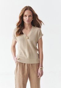 Top de punto beige con escote en V, mangas cortas y dobladillo acanalado. Combinado con pantalones de lino marrón claro y acentos de joyería dorada.