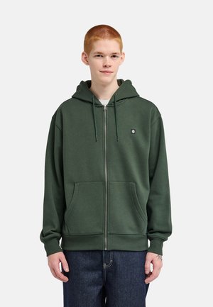 Giovane con capelli rossi corti, indossa una felpa con cappuccio verde scuro con zip e jeans blu scuro, in piedi davanti a uno sfondo semplice.