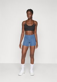 Reggiseno sportivo nero con spalline sottili, abbinato a pantaloncini alti blu, su uno sfondo bianco semplice. Sneakers e calze bianche incluse.