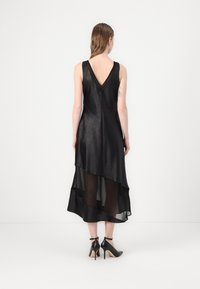 Black sleeveless dress with a V-back, layered design featuring sheer and opaque materials, and a flowing hemline, paired with black high heels.