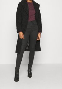 Manteau long noir texturé, porté sur un top violet foncé, associé à un jean taille haute noir et des bottines noires.