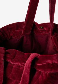 Diep rode fluwelen tote bag met een zachte textuur, brede handvatten en een drukknoopsluiting aan de bovenkant.