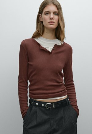 Chemise à manches longues couleur rouille avec un col rond et trois boutons au cou ; portée sur une couche blanche, associée à un pantalon sombre et une ceinture noire.