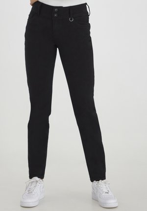 Černé slim-fit kalhoty mají zapínání na dva knoflíky, boční kapsy a poutka na opasek, v kombinaci s bílými teniskami pro kontrast na neutrálním pozadí.