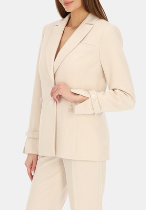 Kobieta ubrana w beżową, dopasowaną marynarkę z pasującymi spodniami, trzymająca jeden guzik prawą ręką, na jednolitym tle.