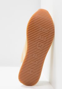 Semelle d'une basket présentant une texture en caoutchouc lisse de couleur orange avec des rainures horizontales et un logo de la marque en relief. Design minimaliste dans un cadre neutre.