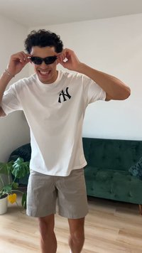 Wit T-shirt met zwart logo, grijze korte broek en zwarte zonnebril. Krullend haar, staand voor een groene fluwelen bank en een potplant.