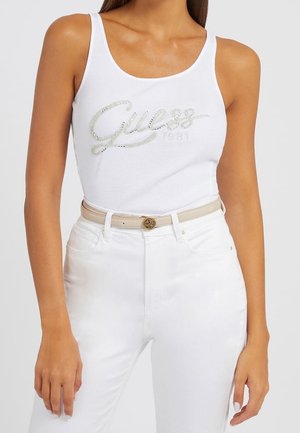 Vrouw die een witte mouwloze tanktop draagt met het 'Guess 1981' rhinestone-logo, witte broek en een beige riem met een ronde gouden gesp.