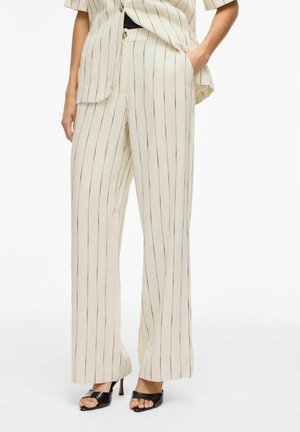 Kvinde iført cremefarvede vidde bukser med tynde lodrette sorte striber, kombineret med matchende kortærmet blazer og sorte hælede sandaler.