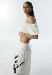 Witte off-the-shoulder top met pofmouwen, gecombineerd met een witte rok met een zwarte handgeschreven zin. Zachte katoenen stof, gladde textuur.