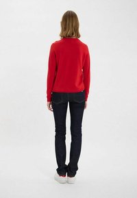 Un pull en maille rouge associé à un jean en denim foncé. Le pull présente un col rond et une coupe ajustée. Des baskets blanches simples complètent le look.