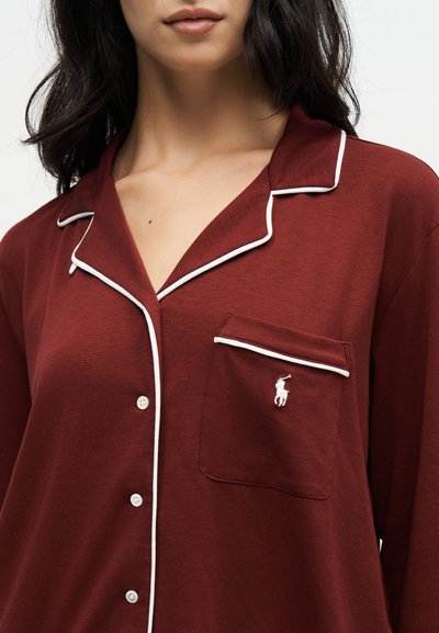 Femme portant une chemise bordeaux boutonnée avec un liseré blanc et un petit logo blanc brodé sur la poche poitrine.
