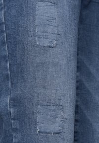 Džínové kalhoty v světle modré barvě s opotřebovanými detaily. Obsahují třepené záplaty na přední straně, které zdůrazňují vybledlý, texturovaný vzhled.