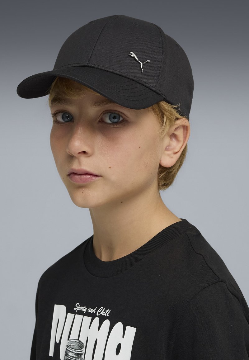 Jeune avec des yeux bleus portant une casquette Puma noire et un t-shirt Puma noir avec un logo blanc sur un fond gris uni.