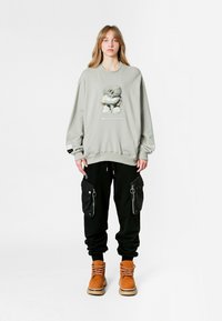 Sudadera gris claro con un diseño gráfico en el centro, combinada con pantalones cargo negros con bolsillos con cremallera y botas naranjas.