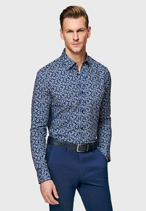 Mężczyzna w dopasowanej, niebieskiej, kwiecistej koszuli z długim rękawem, wsuniętej w granatowe spodnie z czarnym paskiem, stojący na jednolitym tle.