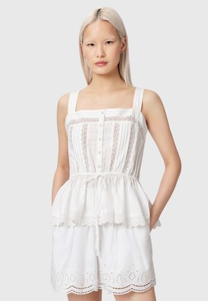 Vrouw met steil blond haar, het dragen van een mouwloze witte jurk met kanten rand, knopen en een trekkoord in de taille, staand tegen een grijze achtergrond.