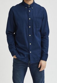 Muž oblečený v tmavě modré košili na knoflíky s dlouhými rukávy a modrých džínách, stojící s jednou rukou v kapse.