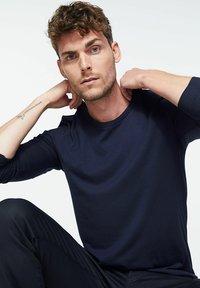 Marineblaue Langarm-T-Shirt aus glattem Material, mit rundem Halsausschnitt und taillierter Passform, kombiniert mit dunklen Hosen. Modell mit zerzaustem Haar posiert lässig.