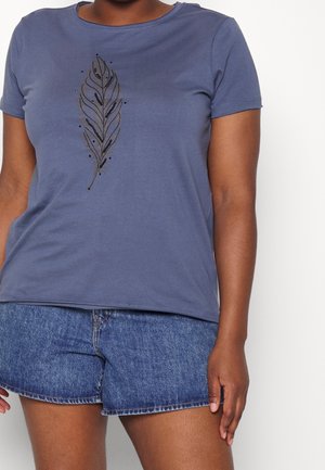 Kobieta ubrana w niebieską koszulkę z krótkim rękawem z czarnym wzorem pióra oraz niebieskie jeansowe szorty, stojąca na białym tle.