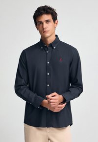 Dunkelblaues Hemd mit Knöpfen aus glattem Stoff, mit einem kleinen roten Logo auf der linken Brust und weißen Knopfakzenten. Lange Ärmel und ein Kragen.