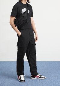 Black T-shirt with a white graphic, black cargo pants with side pockets, and black/red sneakers. Black crossbody bag with textured design.