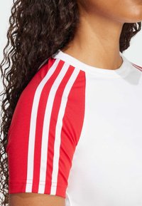 Haut de sport rouge et blanc à manches courtes avec deux rayures blanches sur les épaules rouges, col rond et texture lisse.