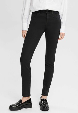 Femme portant un jean noir slim, une chemise blanche à manches longues et des mocassins noirs brillants, debout contre un fond clair uni.