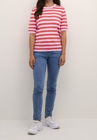 Mujer con camisa de manga corta a rayas rosa y roja, jeans ajustados azules y zapatillas blancas, de pie frente a un fondo claro liso.