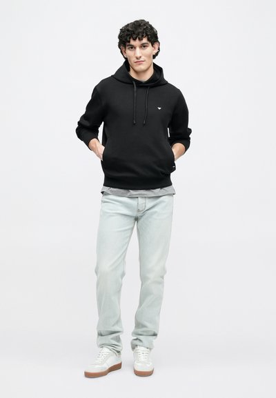 Melna kapuce džemperis ar priekšējo kabatu, gaiši zili džinsi un balti sporta apavi ar brūnām zolēm, attēloti pret vienkāršu fonu.