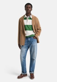 Brauner Wollmischmantel, Polohemd mit grünen und weißen Streifen, hellblaue Jeans, braune Schnürschuhe mit Gummisohle, stehend vor einem weißen Hintergrund.