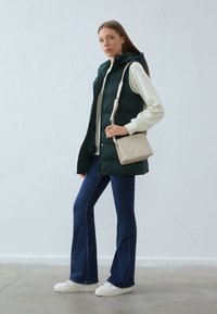 Tamnozeleni zimski prsluk s kapuljačom preko svijetlo kremastog pulovera, uparen s tamnoplavim hlačama s razguženim nogavicama i bijelim tenisicama; drži bež torbicu za crossbody.