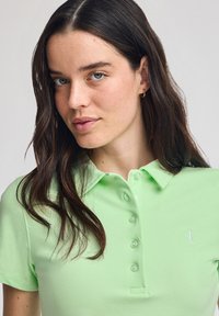 Ljust grönt pikétröja i mjukt tyg, med knappslå och korta ärmar. En liten vit logotyp på vänster bröst är ett framträdande inslag.