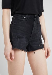Shorts de mezclilla negros de talle alto con dobladillo deshilachado, detalles desgastados y estilo de cinco bolsillos. La tela tiene un ligero estiramiento y un ajuste casual.