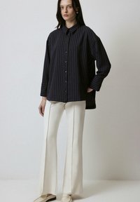 Oversized black striped shirt with buttons, featuring a collar and long sleeves, paired with cream flared trousers. Smooth texture.