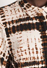 Chemise à manches longues en tissu léger avec un motif abstrait marron et noir sur fond crème, dotée d'un col classique et de boutons.