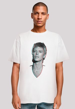 Man wearing white T-shirt with a black-and-white glitch-effect portrait of a person with short hair and a cross necklace, standing against a gray background.