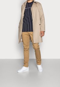 Beige trenchcoat över en marinblå randig t-shirt, khakibyxor och vita sneakers. Kappan är lång med en slät textur och knäppningar.