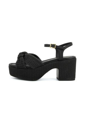 Sandalo platform nero intrecciato con tacco spesso, cinturino alla caviglia con fibbia dorata e dettaglio anteriore annodato.