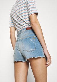 Džíny šortky světle modré barvy s roztrhaným lemem, vybavené koženou nálepkou na pasu a prošívanými detaily na zadních kapsách.