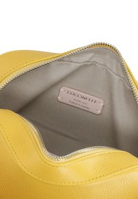 Borsa a tracolla gialla in pelle con chiusura a zip, texture liscia e interno beige con etichetta di branding "Coccinelle."