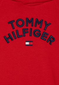 Sweat à capuche rouge avec broderie bleu marine. Présente le texte "TOMMY HILFIGER" en demi-cercle et un petit logo avec des accents rouges, blancs et bleus.