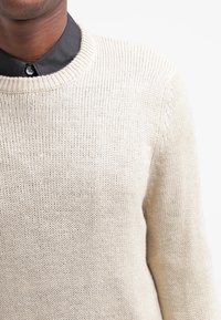 Beżowy sweter dzianinowy z okrągłym dekoltem, charakteryzujący się teksturowanym materiałem i ściągaczami przy rękawach. Noszony na ciemnej koszuli z guzikami.