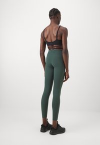 Black sports bra with thin straps, paired with high-waisted green leggings. Model wearing black athletic shoes, viewed from the back.