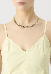 Žena v zlatej hrubej reťazovej náhrdelníku s klipsom zdobeným rhinestone, skombinovaná s svetložltým topom bez rukávov.