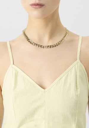 Woman wearing a gold chunky chain necklace with a rhinestone-encrusted clasp, paired with a light yellow sleeveless top.