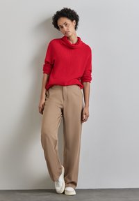 Maglione rosso lavorato a maglia con collo a caramello, abbinato a pantaloni larghi beige e sneaker bianche, creando un look rilassato.