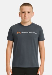 Camiseta gris de manga corta con cuello redondo. Presenta un logo blanco de "UNDER ARMOUR" y un emblema naranja en el lado izquierdo del pecho.