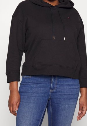 Sudadera corta negra hecha de una tela suave, con un bolsillo delantero, cordones con puntas metálicas, combinada con jeans de color azul oscuro.